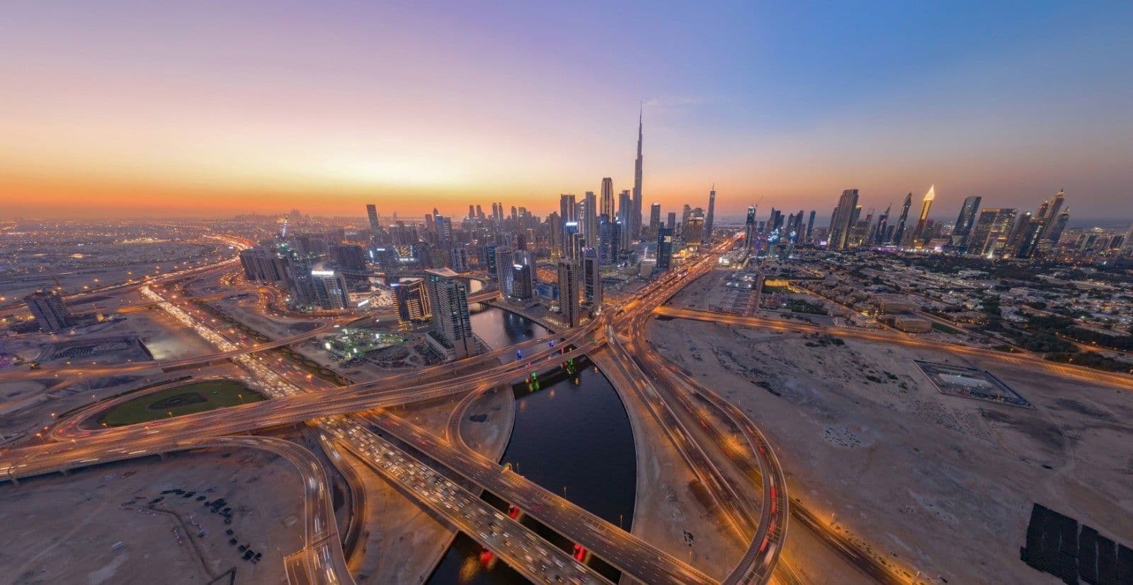 Aerial sunset view of a modern Middle Eastern city with sweeping multi-level highways curving over a canal toward a dense skyline, dominated by a very tall needle-like tower on the horizon.