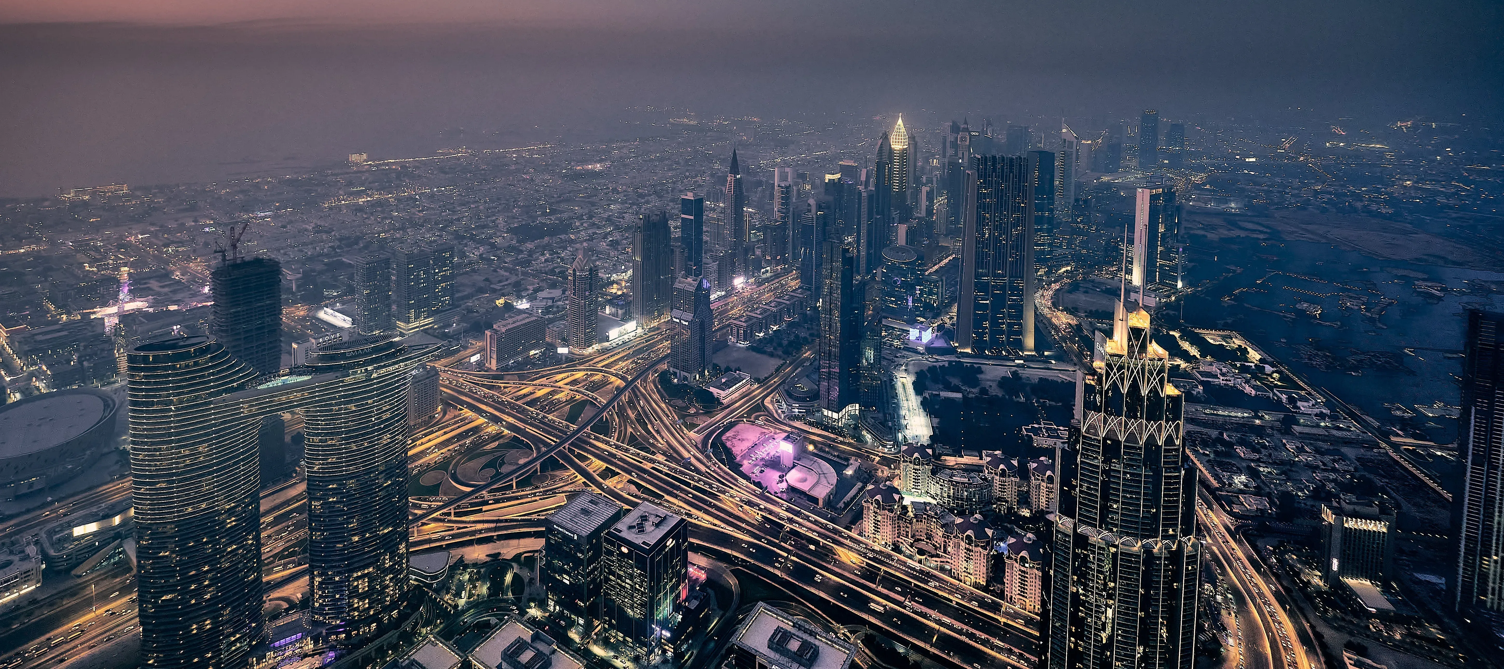 Aerial evening view of Dubai city with brightly lit skyscrapers, intersecting highways, and waterfront in the background.